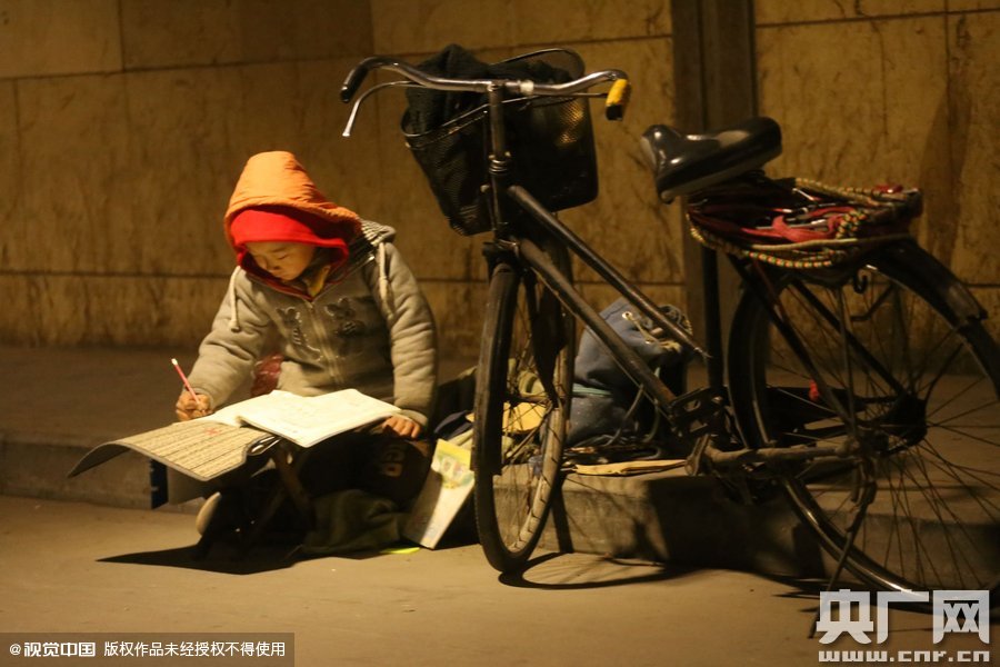 鄭州中原路京廣鐵路大橋下涵洞的馬路牙子邊，一位戴著紅色辣椒帽的小男孩趴在一個小馬扎上，上面鋪著一張車墊，在微弱的燈光下寫作業(yè)