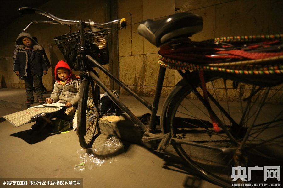 2015年11月8日晚，鄭州，7歲男孩涵洞橋下陪父母練攤，路邊燈下坐小馬扎上寫作業(yè)。