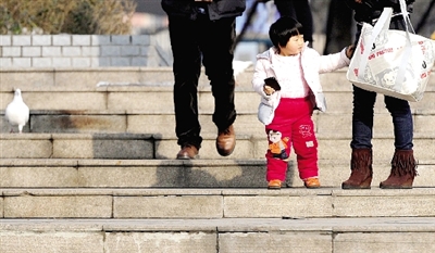 昨天中午，一女孩拉著家長的手在紫荊山公園曬太陽，氣溫升高，孩子的棉衣解開了扣子。